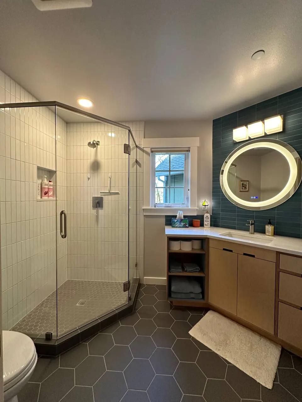 Bathroom with a glass corner shower, black hexagon floor and lit round mirror